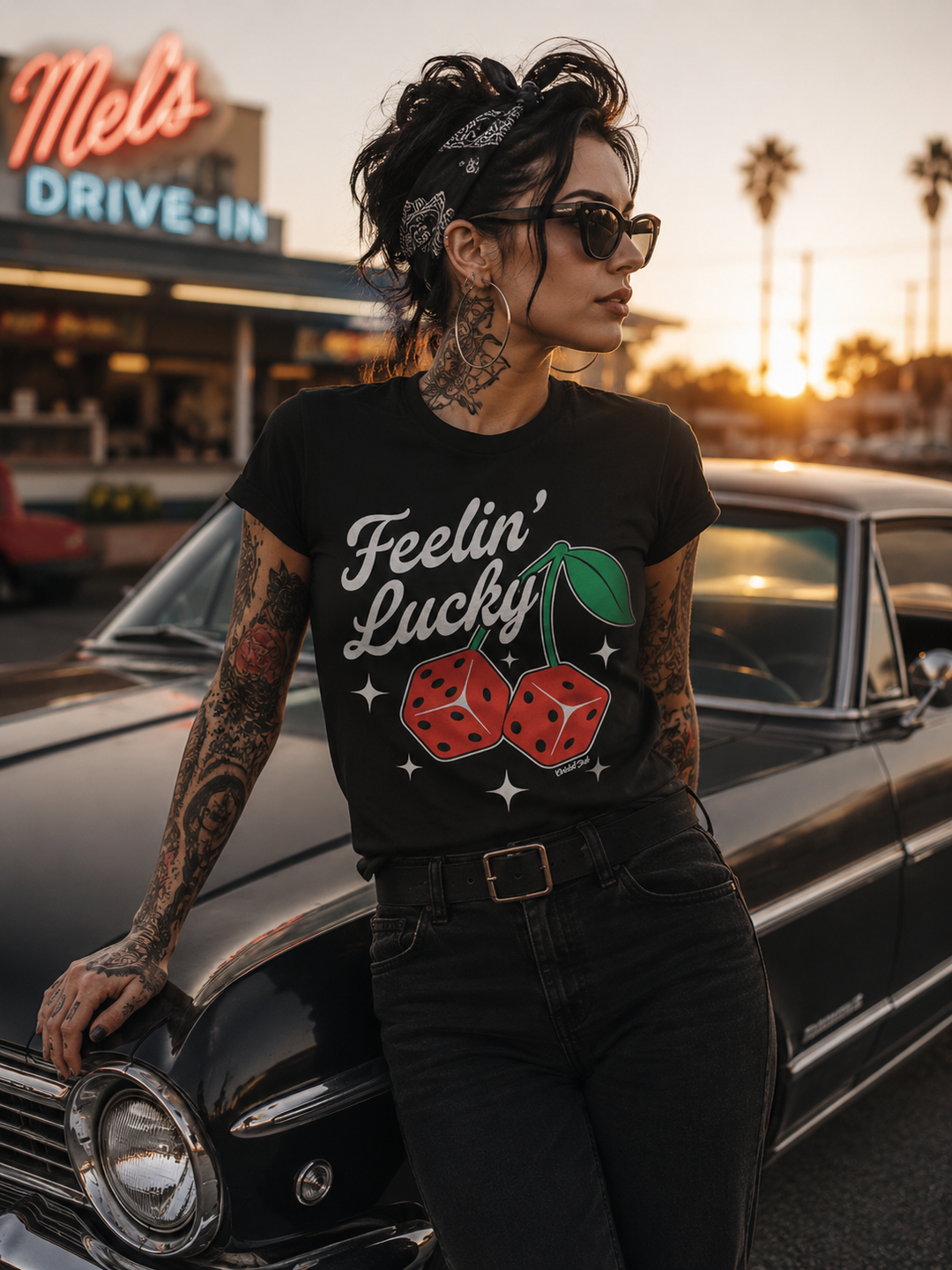 Image of tattooed pin up wearing a black t-shirt with bold feelin lucky print with fun dice that look like cherries, leaning on an old car