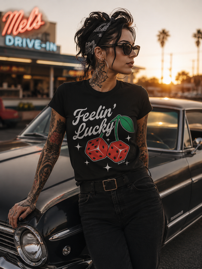 Image of tattooed pin up wearing a black t-shirt with bold feelin lucky print with fun dice that look like cherries, leaning on an old car