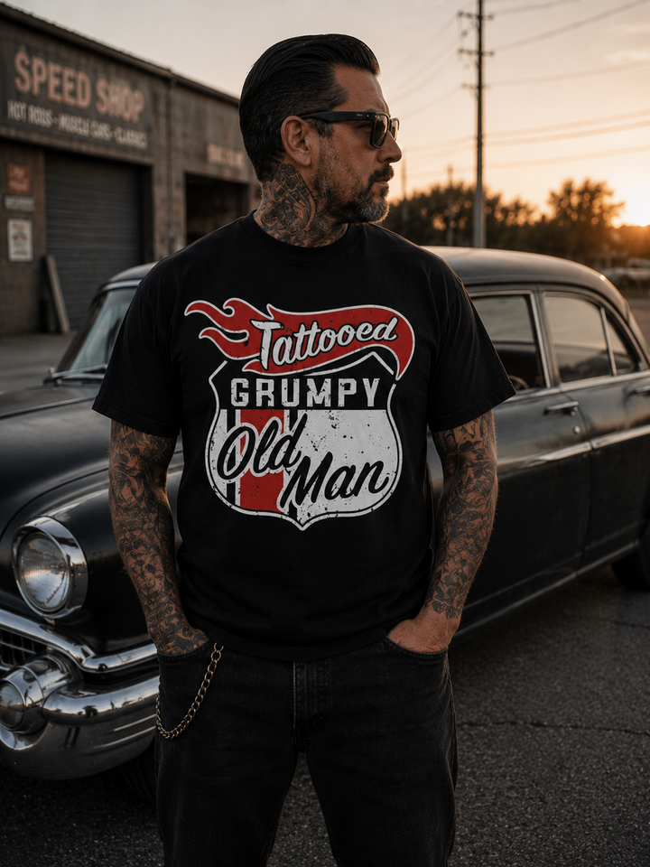 Image of a Tattooed man wearing a black t-shirt with a motive saying "Tattooed Grumpy Old Man" standing in front of an old car