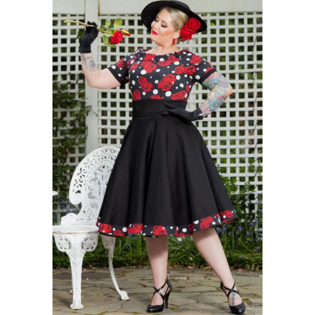 Image of a lady wearing a black dress with rose and white polka dot pattern holding a rose and posing for the camera