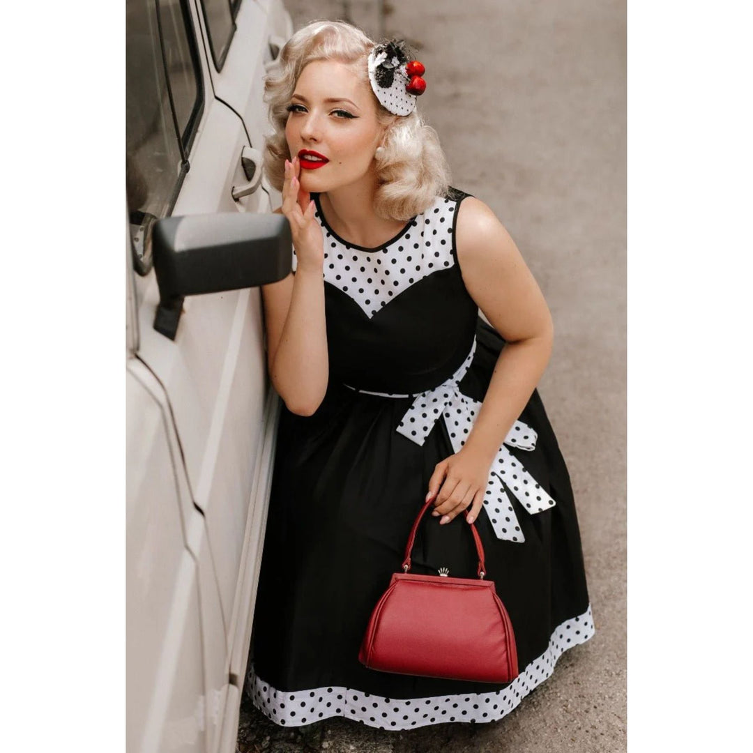 Image of a blonde lady wearing a black dress with white with black polka dot hem and neckline fixing her lipstick in the side mirror of a car