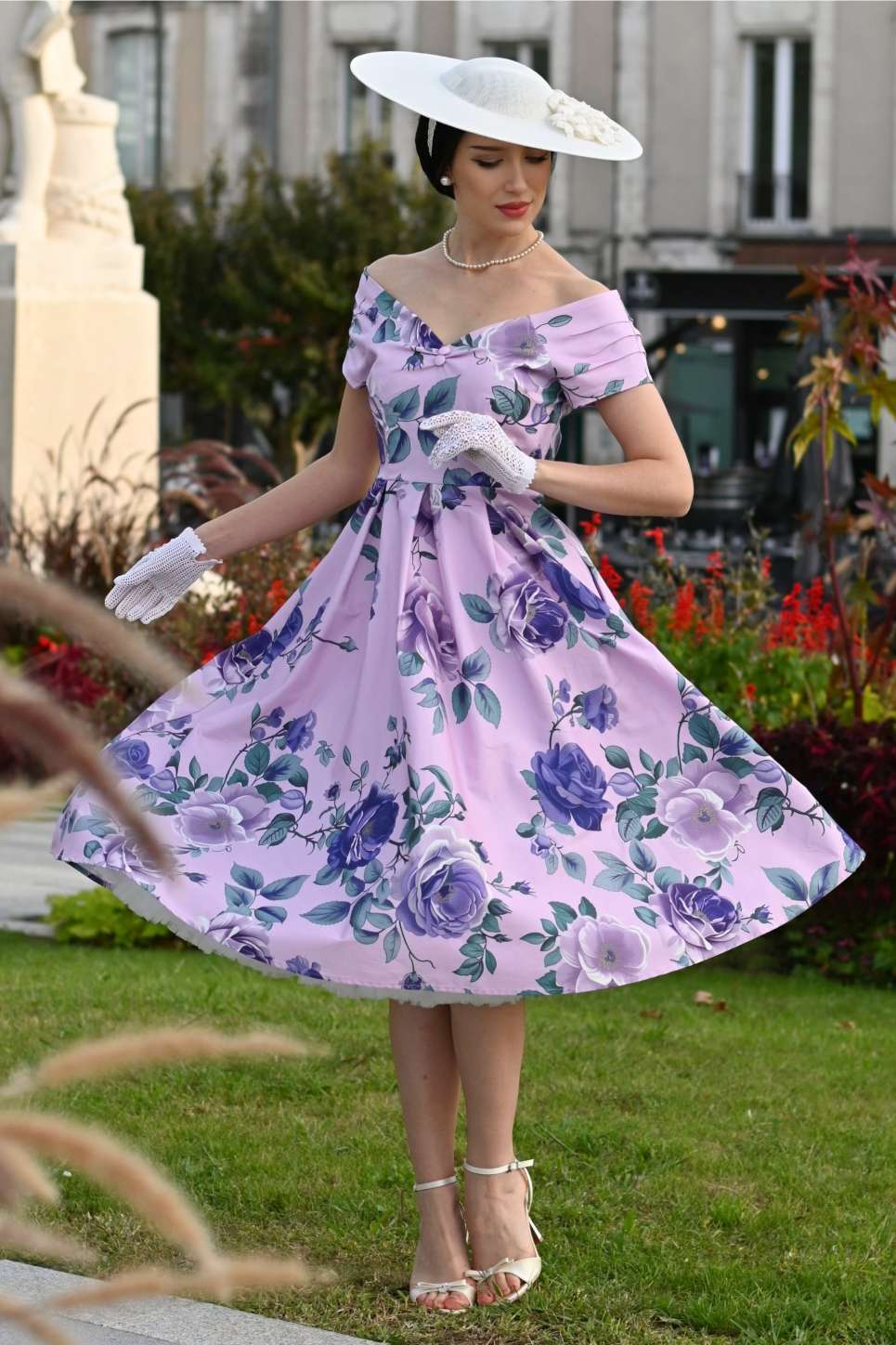 Woman in a purple floral dress standing outdoors with a decorative hat and gloves.