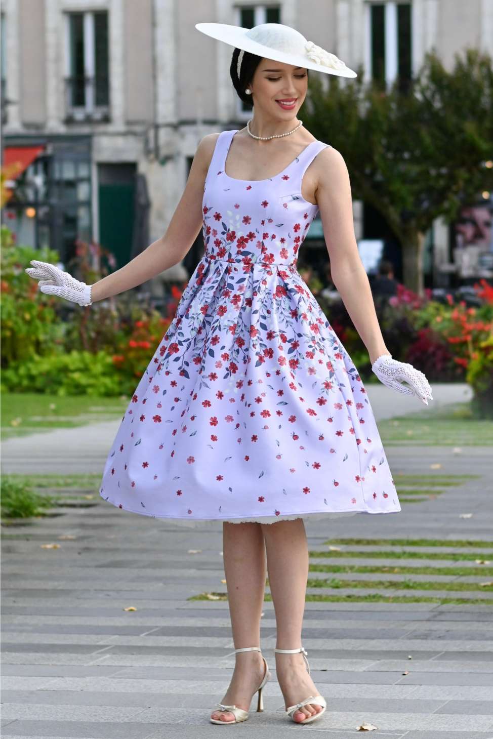 Woman in a floral dress with a white hat and gloves standing on a city street.