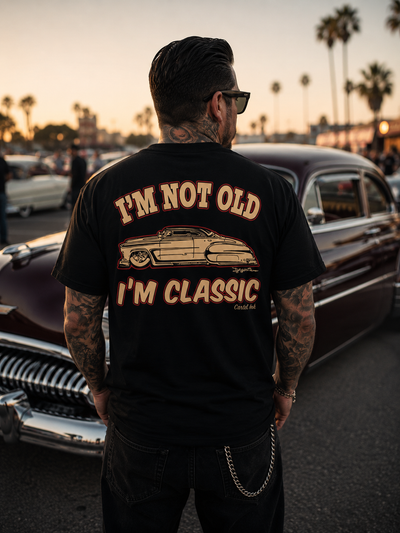Image of a tattooed man standing facing an old car wearing a black t-shirt with I'm not old, I'm classic in bold font on the back