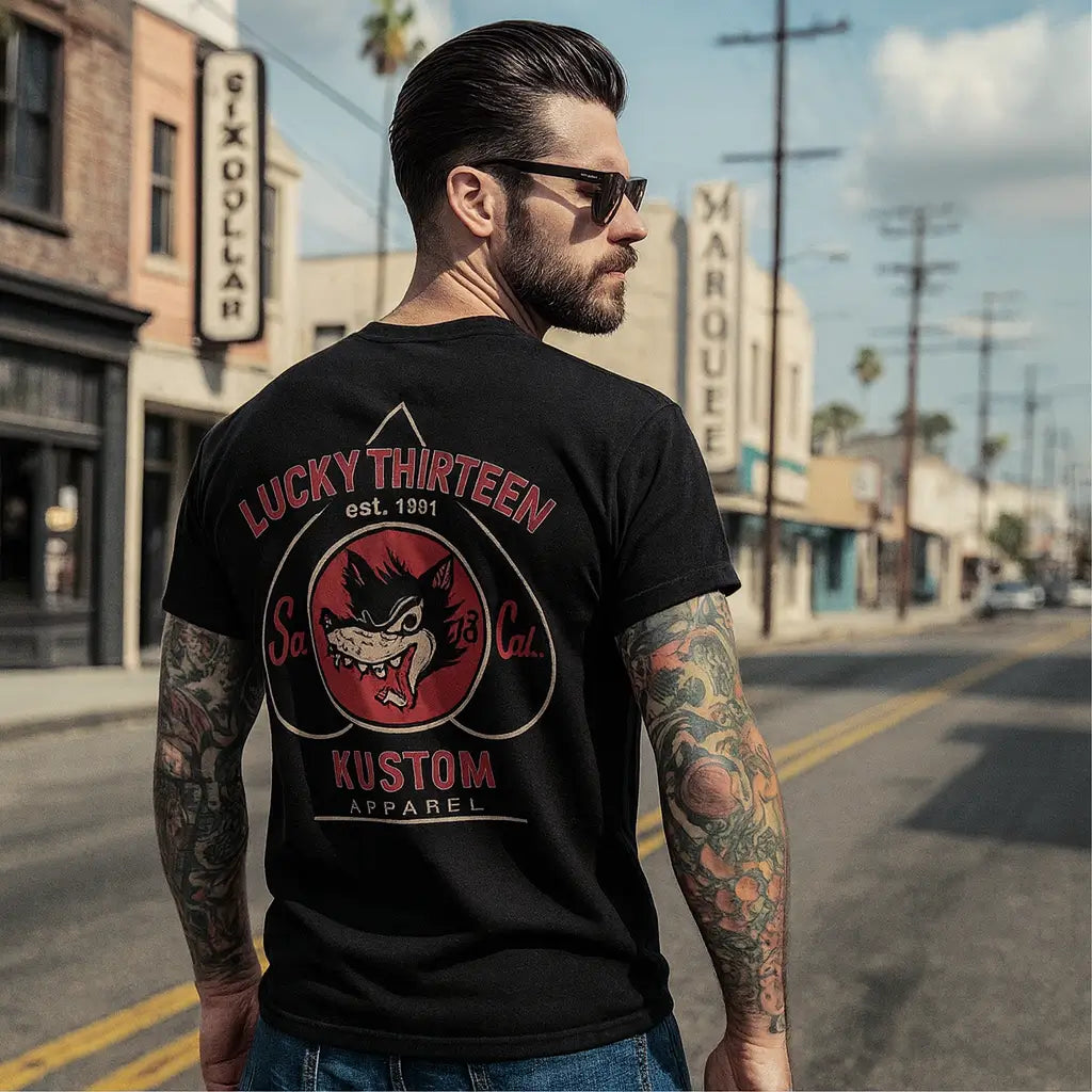 Back view of a man with sleeve tattoos wearing a black Lucky 13 Kustom Apparel t-shirt with a red wolf graphic on a city street.