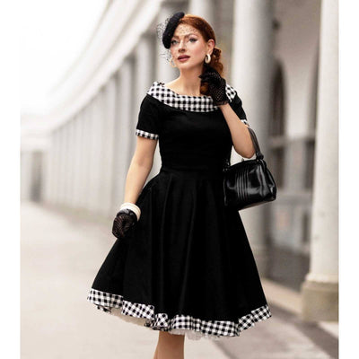 Stunning red haired model wearing the Darlene dress which is black with a black and white gingham timr on the collar, hem and sleeves. She looks very elegant with a hat and bag and gloves